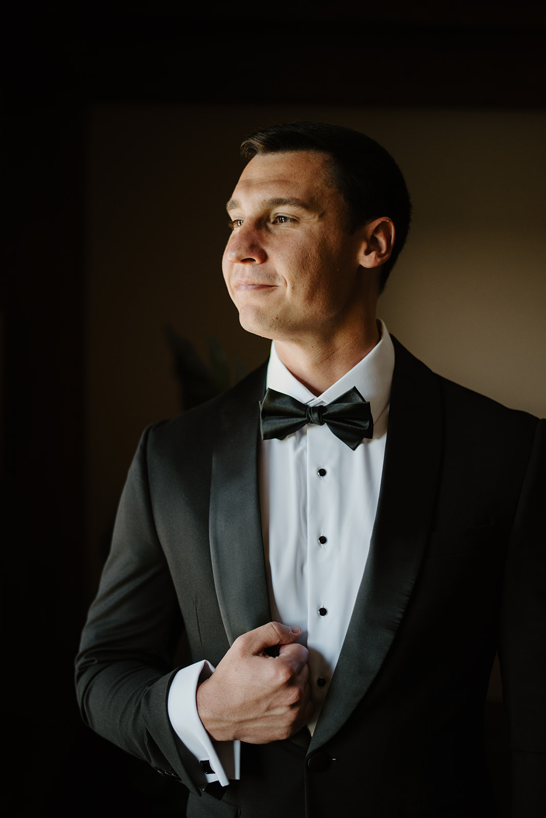 Groom in black tuxedo smiling warmly while looking out the window before his wedding ceremony.